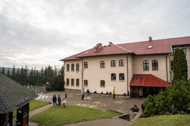 Fotografia przedstawiająca tylną część budynku, z żółtą elewacją. Przed szkołą znajduje się plac z kostki chodnikowej oraz trawniki i zazielenienia.