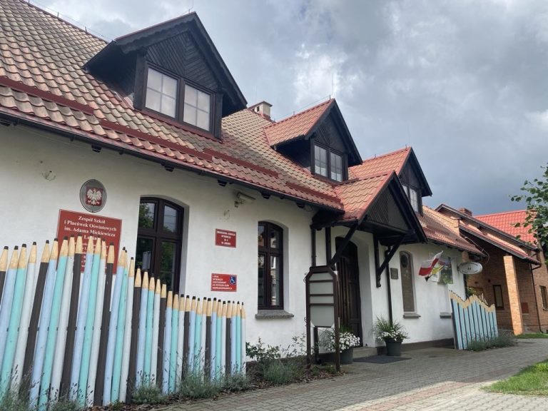 Fotografia przedstawiająca frontową część budynku szkoły, z widocznym wejściem i tablicami informacyjnymi. Elewacja jest w kolorze białym, natomiast dach pokryto czerwonymi dachówkami.