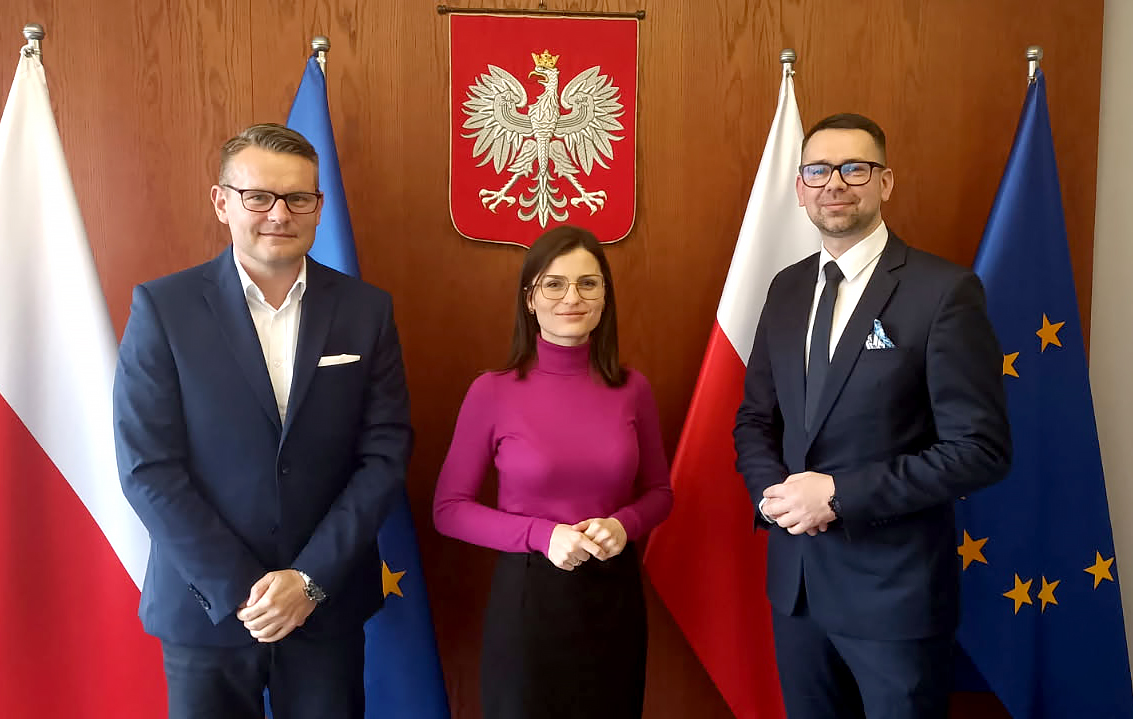 Zdjęcie przedstawia trzy osoby stojące na tle drewnianej ściany z godłem Polski, pomiędzy flagami Polski i Unii Europejskiej. Po lewej stronie znajduje się mężczyzna w granatowym garniturze, białej koszuli i okularach. W środku kobieta w okularach, ubrana w fioletowy golf i czarną spódnicę. Po prawej stronie stoi mężczyzna w ciemnym garniturze, białej koszuli, czarnej krawacie i okularach, z przypiętą niebieską wstążką do klapy marynarki. Wszyscy patrzą na aparat i mają neutralne wyrazy twarzy.