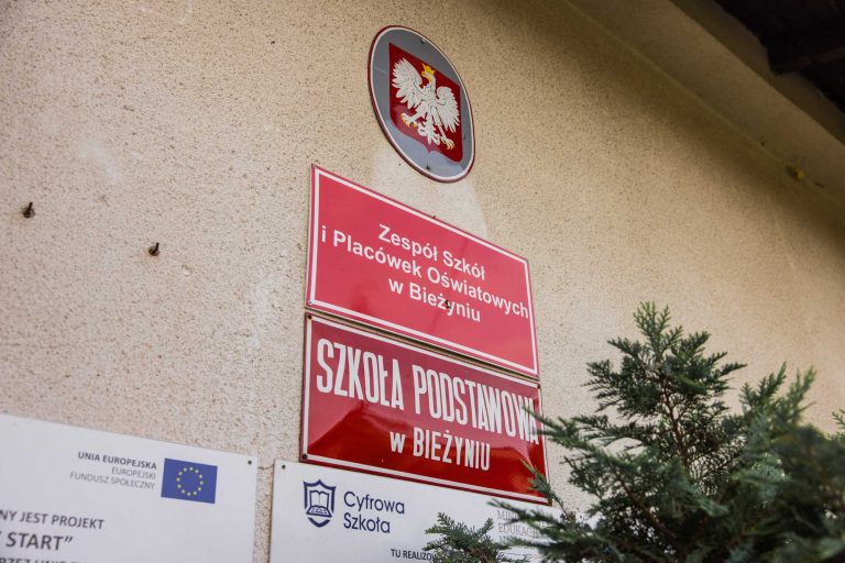 Fotografia przedstawiająca budynek szkoły, ze skupieniem na przedniej części nieruchomości, w szczególności tablic informujących o przeznaczeniu budynku.