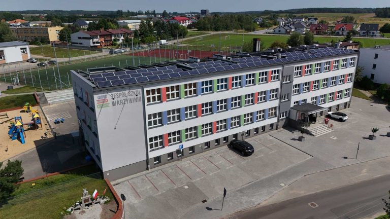 Fotografia z drona przedstawiająca gmach budynku. Na dachu zainstalowane są panele fotowoltaiczne. Elewacja jest w przeważająco biały kolorze, ze wstawkami w kolorze niebieskim, zielonym i czerwonym. Przed budynkiem znajduje się parking.