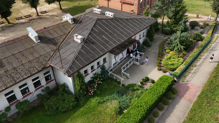 Fotografia z drona przedstawiająca budynek szkoły, ze skupieniem na przedniej części nieruchomości, z wyraźnie widoczną rampą podjazdową dla niepełnoprawnych oraz czarnym dachem i elewacją w jasnym kolorze.