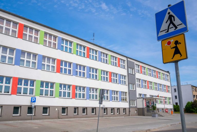 Fotografia przedstawiająca gmach budynku. Na dachu zainstalowane są panele fotowoltaiczne. Elewacja jest w przeważająco biały kolorze, ze wstawkami w kolorze niebieskim, zielonym i czerwonym. Przed budynkiem znajduje się parking.