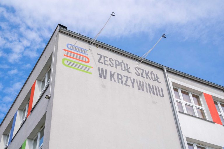 Fotografia przedstawiająca gmach budynku. Na dachu zainstalowane są panele fotowoltaiczne. Elewacja jest w przeważająco biały kolorze, ze wstawkami w kolorze niebieskim, zielonym i czerwonym. Przed budynkiem znajduje się parking.