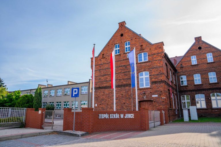 Fotografia przedstawiająca ceglany budynek szkoły. Na pierwszym planie widać flagi, w tym Polski oraz UE oraz napis Zespół Szkół w Jerce