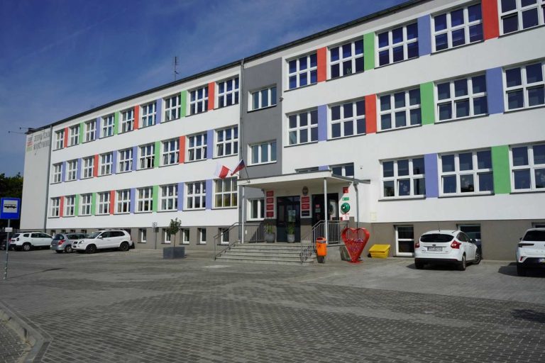 Fotografia przedstawiająca gmach budynku. Na dachu zainstalowane są panele fotowoltaiczne. Elewacja jest w przeważająco biały kolorze, ze wstawkami w kolorze niebieskim, zielonym i czerwonym. Przed budynkiem znajduje się parking.