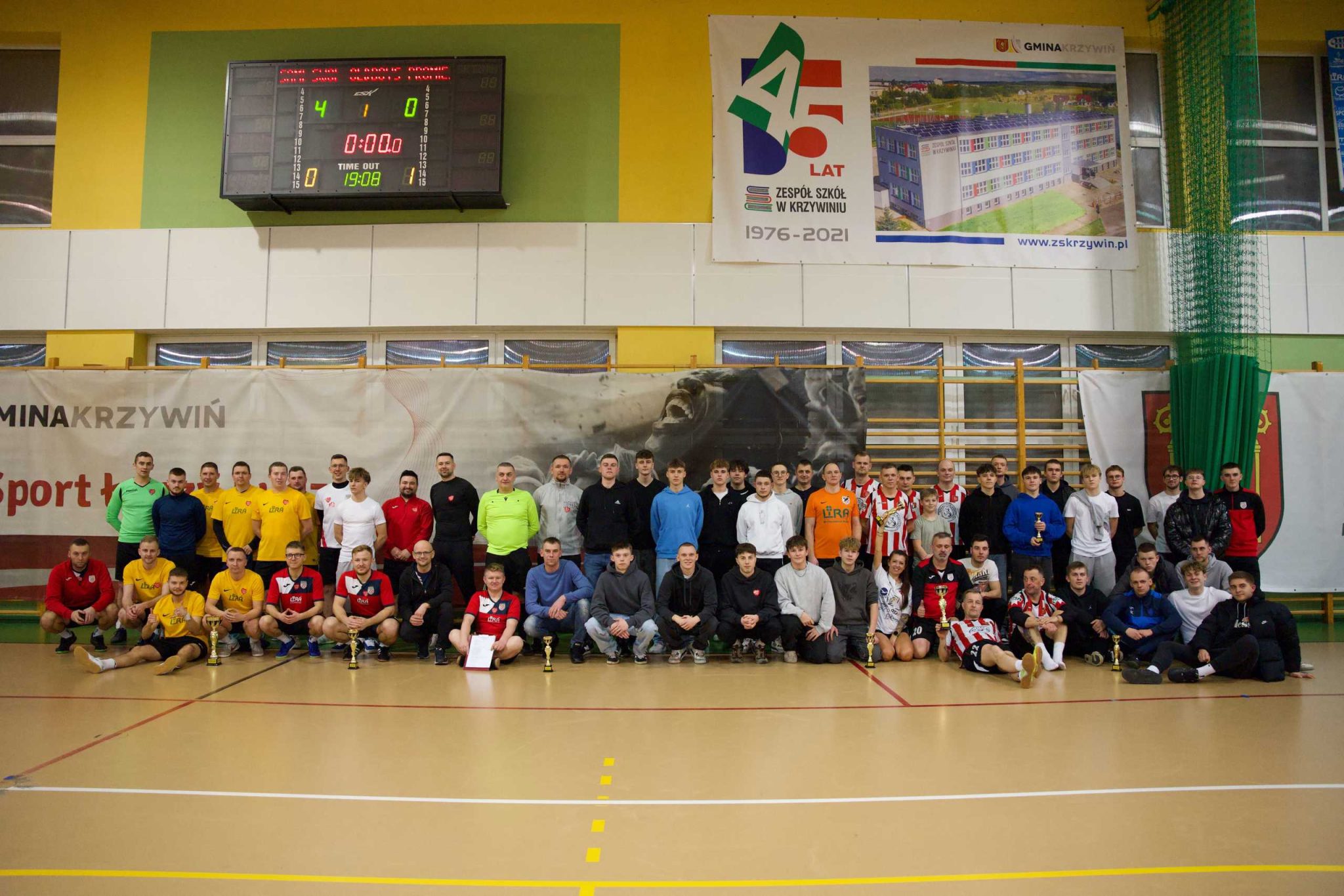 Zdjęcie grupowe uczestników turnieju piłki nożnej halowej w hali sportowej w Krzywiniu. Na pierwszym planie zawodnicy w kolorowych strojach sportowych, niektórzy trzymają puchary i dyplomy. W tle tablica wyników pokazująca wynik 4:0, banery promocyjne Gminy Krzywiń oraz Zespołu Szkół w Krzywiniu. Grupa składa się z młodzieży, trenerów i dorosłych zawodników, wszyscy ustawieni do wspólnej fotografii po zakończeniu rozgrywek