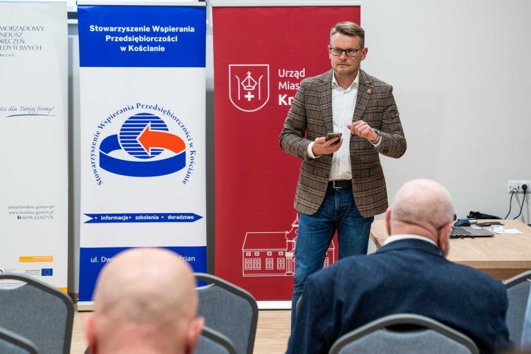 Zdjęcie przedstawia mężczyznę w okularach, ubranego w brązową marynarkę, białą koszulę i jeansy, który prowadzi prezentację, trzymając telefon w dłoni. W tle znajdują się banery promujące Stowarzyszenie Wspierania Przedsiębiorczości w Kościanie oraz Urząd Miasta i Gminy Krzywiń. Na pierwszym planie widać uczestników spotkania siedzących na krzesłach, w tym dwóch mężczyzn z łysymi głowami, zwróconych w stronę prelegenta. Po prawej stronie znajduje się stół z laptopem, filiżanką oraz notatkami.
