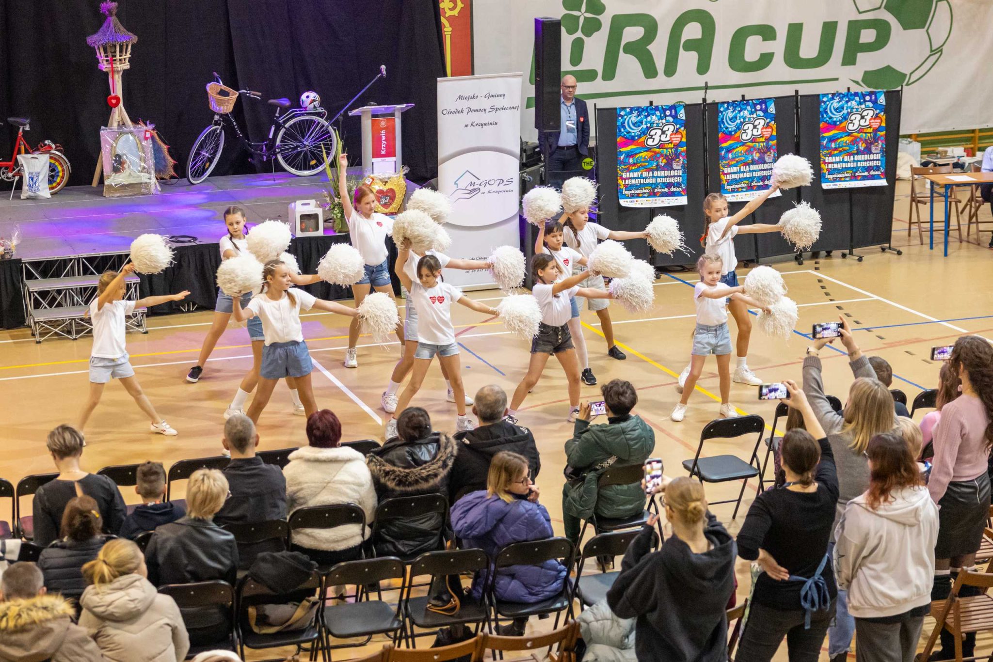 Zdjęcie przedstawia występ dziecięcej grupy tanecznej na hali sportowej. Dziewczynki ubrane w białe koszulki i krótkie spodenki prezentują układ choreograficzny z białymi pomponami. W tle widoczna jest scena z dekoracjami, w tym rowerami i rekwizytami, oraz baner Miejsko-Gminnego Ośrodka Pomocy Społecznej w Krzywiniu. Na trybunach siedzą widzowie, z których wielu nagrywa występ telefonami. Po prawej stronie widać banery promujące 33. Finał Wielkiej Orkiestry Świątecznej Pomocy, a na ścianie widnieje napis 'IRA CUP