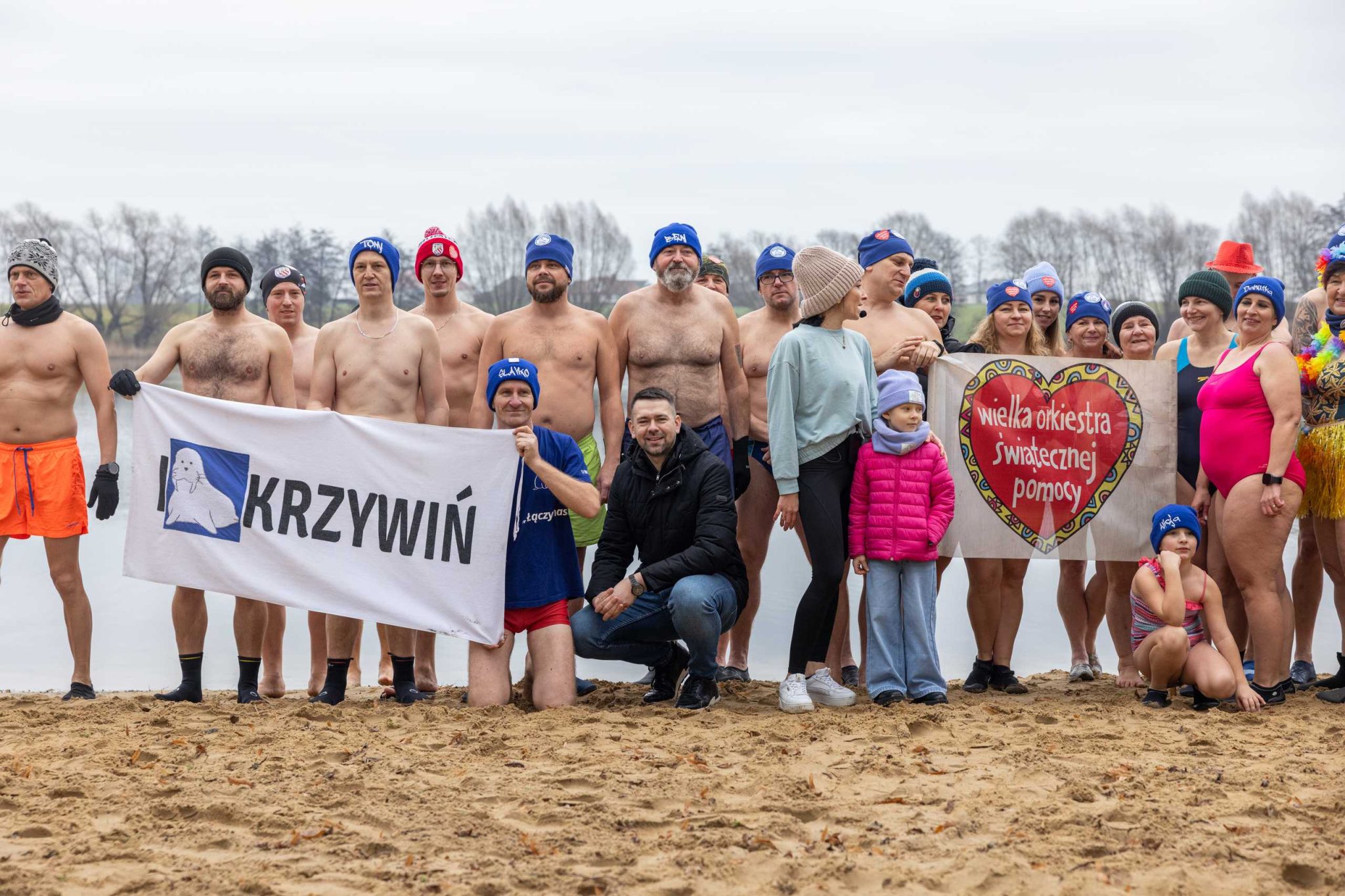 Grupa morsów na plaży nad jeziorem podczas zimowej kąpieli, ubrana w stroje kąpielowe i czapki. Część osób trzyma baner z napisem 'KRZYWIŃ' i logo morsów, a inni trzymają materiał z sercem i napisem 'Wielka Orkiestra Świątecznej Pomocy'. W tle widać jezioro i bezlistne drzewa, a na piasku stoi także kilkoro dzieci w zimowych ubraniach. Atmosfera radosna, uczestnicy uśmiechnięci, gotowi do zimowej kąpieli.