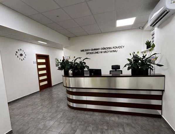 Fotografia przedstawia nowoczesną recepcję MGOPS w Krzywiniu. Pomieszczenie utrzymane jest w jasnych kolorach. W centralnym punkcie umiejscowiona drewniana lada zdobiona metalowymi, srebrnymi pasami. Na podłodze położone są płytki.