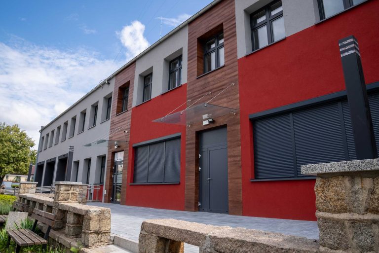 Zdjęcie przedstawia fragment budynku Ochotniczej Straży Pożarnej w Krzywiniu. Budynek rozciąga się wzdłuż kadru, dominujące kolory to szary i czerwony z drewnianymi uzupełnieniami w miejscach gdzie są drzwi.