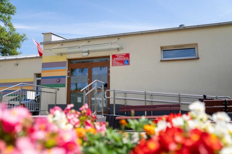 Budynek z wejściem, rampą dla niepełnosprawnych, tabliczką "Obwodowa Komisja Wyborcza nr 18" i flagą Polski, z kwiatami na pierwszym planie.