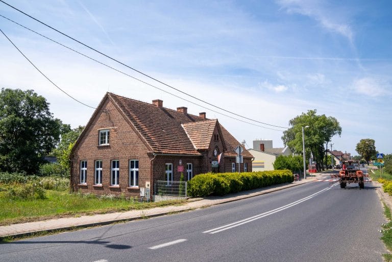 Ceglany budynek z dwuspadowym dachem, w którym mieści się Zespół Szkół i Placówek Oświatowych im. Adama Mickiewicza w Lubieniu oraz Przedszkole Samorządowe w Bielewie, zlokalizowany przy drodze w Bielewie, z widocznym traktorem w oddali.