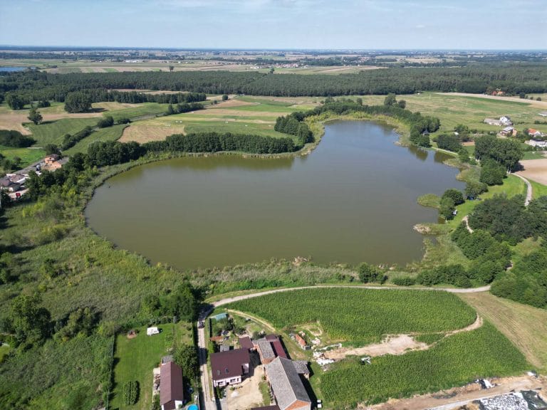 Fotografia lotnicza przedstawiająca jezioro Bieżyń. Otoczone jest licznymi drzewami oraz łąką. Przy jeziorze znajdują się zabudowania.