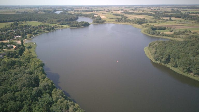 Fotografia lotnicza przedstawiająca jezioro Cichowo. Widać liczne drzewa oraz jezioro Mórka na horyzoncie. Na wodzie można zauważyć kilka pływających obiektów przypominających łódki lub rowery wodne.