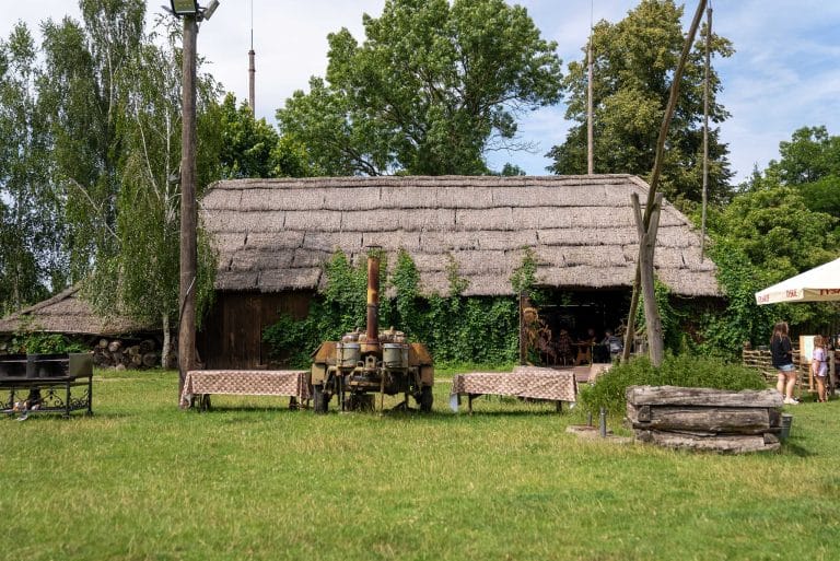 Wiejska chata z krytym strzechą dachem. Przed chatą stoją dwa stoły a pomiędzy nimi wojskowa kuchnia polowa z grochówką. Po prawej stronie od stołów widać ciemną drewnianą studnię, a po lewej kawałek grilla.