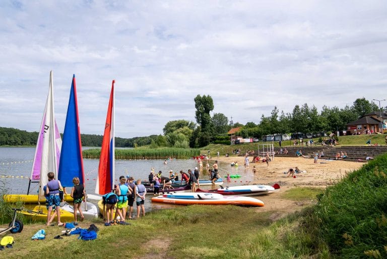 Zdjęcie przedstawia plażę w Cichowie. Na pierwszym planie widać kilka grup osób które przygotowują się do wypłynięcia łodziami. Na kolejnych planach można zauważyć liczne grupy osób które pływają, lub siedzą na plaży. W oddali widać zabudowania oraz dużą ilość zieleni.