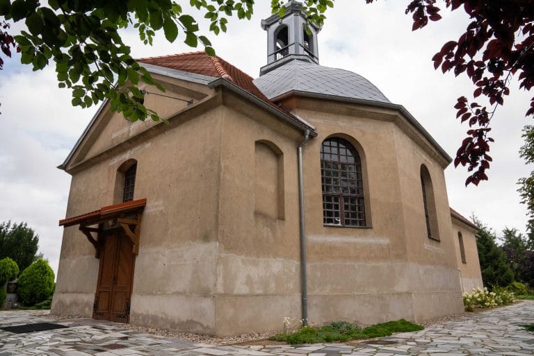 Zdjęcie przedstawia kościół Rzymskokatolicki pw. św. Idziego widziany z boku. Można zauważyć apsydę z oknami i szarym dachem który przechodzi w spadowy czerwony dach. Na apsydzie jest szara wieżyczka z dzwonem. Bezpośrednio nad frontowymi drzwiami widać mały czerwony daszek.