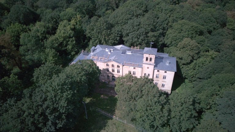 Fotografia lotnicza przedstawiająca pałac w Czerwonej Wsi. Jest on otulony drzewami. Ma trzy piętra oraz wieżyczkę która jest najwyższym punktem w budynku; na jej szczycie widać wiatrowskaz.