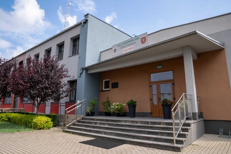 Zdjęcie przedstawia fragment budynku Ochotniczej Straży Pożarnej w Krzywiniu. Widać wejście do sali widowiskowej. Nad wejściem znajduje się tablica z napisem "SALA WIDOWISKOWA DOM STRAŻAKA KRZYWIŃ". Do wejścia prowadzą schody. Przed budynkiem rosną drzewa i krzewy.