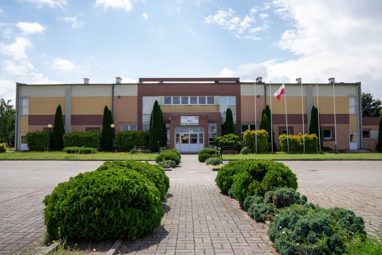 Zdjęcie przedstawia halę sportowo-widowiskową w Krzywiniu. Jest to różno-kolorowy prosty budynek. Przed budynkiem wisi flaga Polski a teren jest porośnięty zielenią. Na pierwszym planie widać parking wyłożony kostką brukową z krzewami pomiędzy.