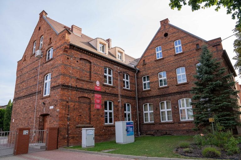 Zdjęcie przedstawia budynek Zespołu Szkół w Jerce z ciemnoczerwonej cegły z asymetryczną bryłą, stromymi dachami, białymi oknami i elementami dekoracyjnymi z cegły. Przed budynkiem trawnik, krzewy, drzewo iglaste i metalowe ogrodzenie.