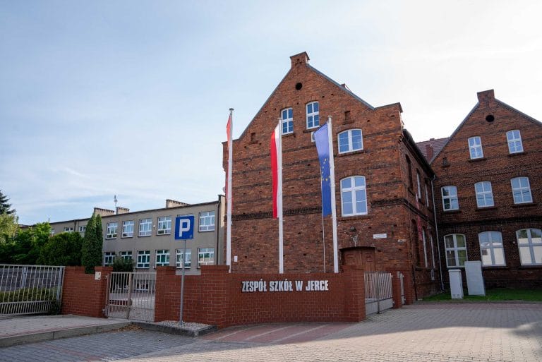 Zdjęcie przedstawia budynek Zespołu Szkół w Jerce z czerwonej cegły, z dwuspadowymi dachami i białymi oknami, z niskim murem z napisem "ZESPÓŁ SZKÓŁ W JERCE" i trzema masztami z flagami. W tle widoczny szary budynek boczny i zieleń.