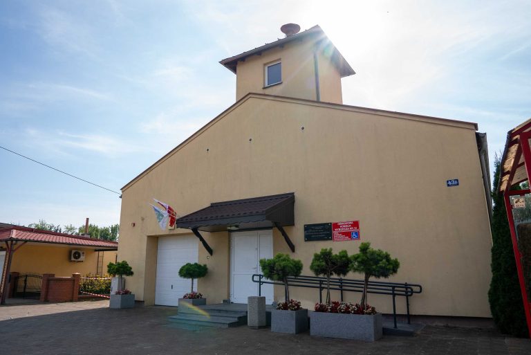 Budynek Ochotniczej Straży Pożarnej w Jurkowie z jasnobeżową elewacją, dwuspadowym dachem i wieżyczką, z flagą, tablicą informacyjną i numerem budynku. Przed wejściem schody, rampa dla niepełnosprawnych, donice z kwiatami i ozdobne krzewy.