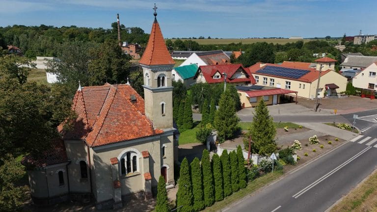 Fotografia lotnicza przedstawiająca kościół rzymskokatolicki w Jurkowie z jasnymi ścianami i czerwonym dachem, z wysoką wieżą i otaczającą zielenią. W tle wiejska zabudowa i pola uprawne. Obok kościoła przebiega asfaltowa droga.