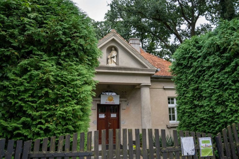 Zdjęcie przedstawia budynek Przedszkola w Kopaszewie z jasną elewacją i czerwonym dachem, z figurą Maryi nad wejściem i tabliczką z napisem "Słoneczka". Budynek jest otoczony zielenią.