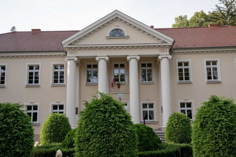 Zdjęcie przedstawia pałac w Kopaszewie, dawna rezydencja rodziny Chłapowskich, z klasycystyczną fasadą, portykiem kolumnowym i datami "1801" i "1923" nad skrajnymi kolumnami. Na fasadzie widać liczne okna oraz czerwony spadowy dach. Otoczony zadbanym ogrodem z formowanymi krzewami.