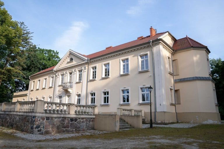 Zdjęcie przedstawia pałac w Kopaszewie z jasną elewacją, czerwonym dachem, centralnym ryzalitem z balkonem i okrągłą wieżą boczną, otoczony zielenią i z kamiennym murem oporowym na pierwszym planie.