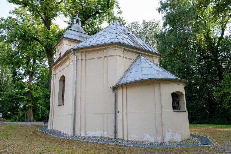 Zdjęcie przedstawia małą, zabytkową kapliczkę o kremowych ścianach i szarym, wielospadowym dachu, z zaokrąglonym prezbiterium i małą wieżyczką, otoczona drzewami.