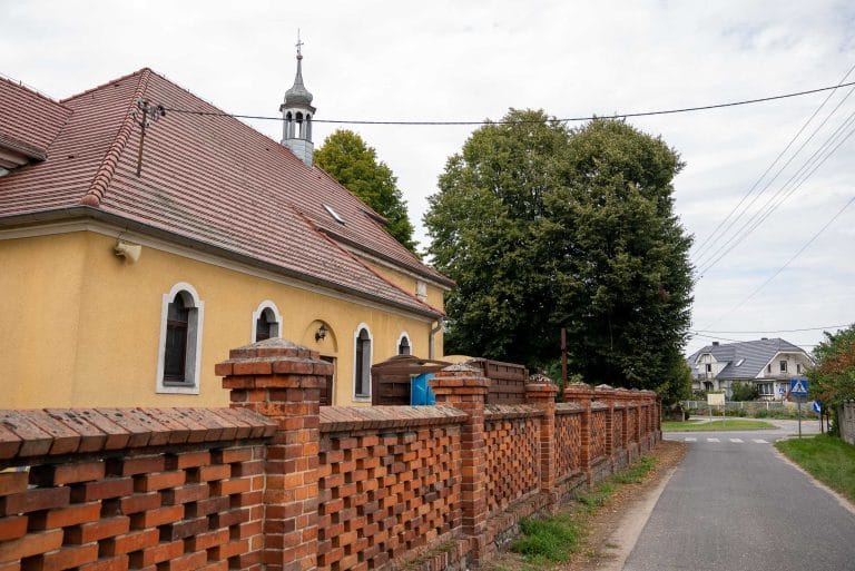Zdjęcie przedstawia fragment kościoła w Kopaszwie z żółtą ścianą i czerwoną dachówką, z małą wieżyczką, oraz mur z czerwonej cegły, widziane z perspektywy ulicy. W tle widoczne domy i zieleń.