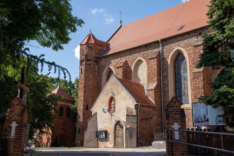 Zdjęcie przedstawia kościół pw. św. Mikołaja Biskupa w Krzywiniu z czerwonym spadowym dachem i charakterystyczną wieżą. Po lewej stronie znajduje się mniejsza, również ceglana budowla. Nad wejściem do kościoła widać figurkę Maryi. Całość otoczona jest zielenią.