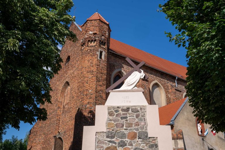 Zdjęcie przedstawia kościół pw. św. Mikołaja Biskupa w Krzywiniu. Jest zbudowany z czerwonej cegły, z charakterystyczną wieżą i czerwonym spadowym dachem. Na pierwszym planie, przed kościołem, stoi pomnik Chrystusa dźwigającego krzyż. Pomnik jest biały, umieszczony na kamiennym cokole.