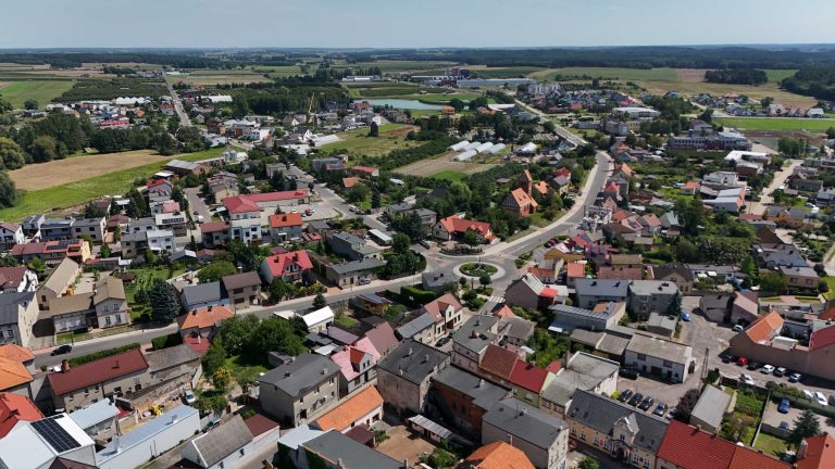 Fotografia lotnicza przedstawiająca Krzywiń. Widać rondo w centrum, otoczone polami i łąkami. Widoczna zwarta zabudowa z domami jednorodzinnymi i siatka ulic.
