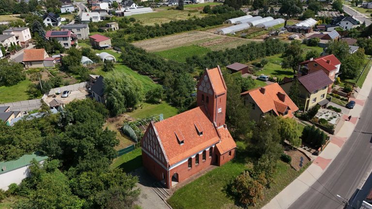 Fotografia lotnicza przedstawiająca kościół poewangelicki w Krzywiniu z czerwonej cegły z masywną wieżą, otoczony zabudową miejską, zielenią i polami uprawnymi. Widoczne drogi i szklarnie.