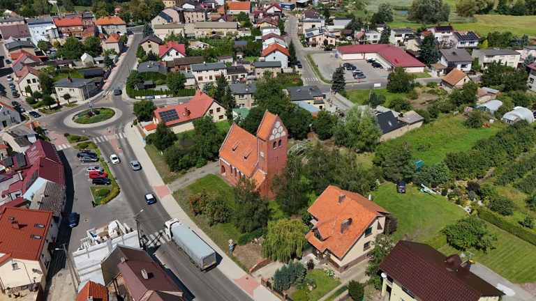 Fotografia lotnicza przedstawiająca Krzywiń. Widoczny kościół poewangelicki z czerwonej cegły z wysoką wieżą w centrum. Jest otoczony zabudową mieszkalną oraz zielenią. Kościół znajduje się przy głównej ulicy w Krzywiniu.