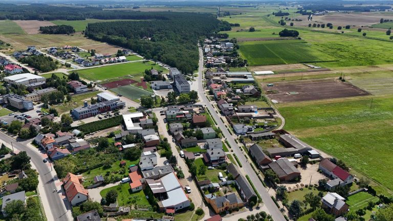 Fotografia lotnicza przedstawiająca Krzywiń. Można zauważyć zróżnicowaną zabudowę, miasto otoczone jest łąkami i polami a na horyzoncie widać gęsty las.