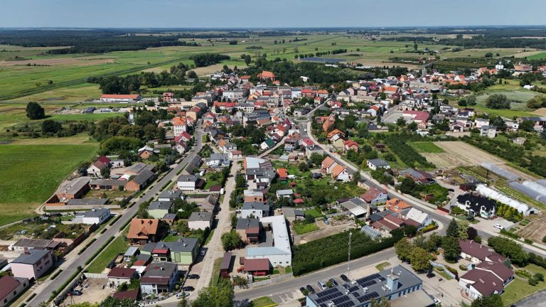 Fotografia lotnicza przedstawiająca Krzywiń. Widać zwartą zabudową domów jednorodzinnych i kilkoma budynkami wielorodzinnymi, z rynkiem w centrum. Widoczna regularna siatka ulic. Miejscowość otoczona jest polami uprawnymi, łąkami i niewielkimi lasami liściastymi.