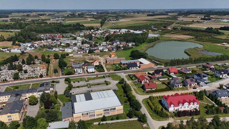 Fotografia lotnicza przedstawiająca Krzywiń. W oczy rzuca się jezioro z małym zbiornikiem retencyjnym obok. Widać gęstą zabudowę z wyróżniającą się Halą Sportowo-Widowiskową. Miasto jest otoczone licznymi drzewami, łąkami i polami.