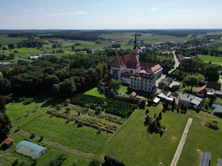Fotografia lotnicza przedstawiająca Opactwo Benedyktynów w Lubiniu. Budynek ten jest wysoki z biała elewacją i czerwonym, spadzistym dachem z wysoką wieżą. Obok, na łącę można zauważyć labirynt z żywo płotu. Cały teren otoczony jest zielenią a przy samym kompleksie jest mały lasek lub park.