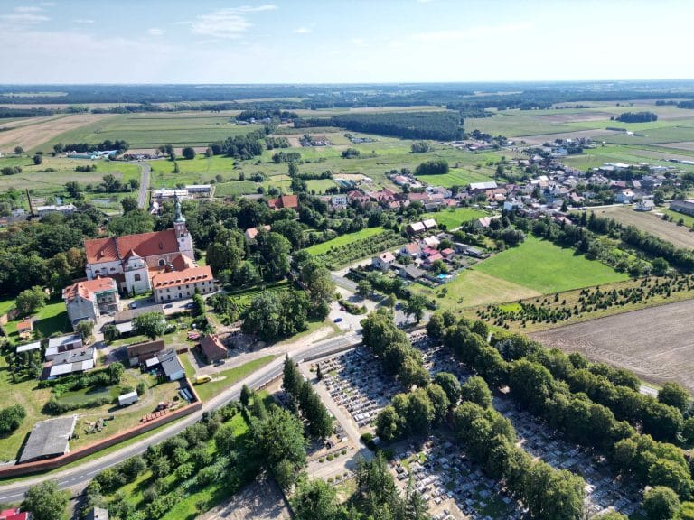 Fotografia lotnicza przedstawiająca panoramę wsi Lubiń. W oczy rzuca się budynek Opactwa Benedyktynów w Lubiniu z przyległym cmentarzem. Można zauważyć dużą ilość zieleni oraz łąk i pól uprawnych. Widoczna jest siatka zabudowań miejskich.