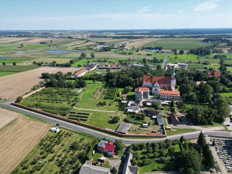 Fotografia lotnicza przedstawiająca panoramę wsi Lubiń. W oczy rzuca się budynek Opactwa Benedyktynów w Lubiniu z labiryntem z żywopłotu. Można zauważyć dużą ilość zieleni oraz łąk i pól uprawnych. Widoczna jest siatka zabudowań miejskich a w oddali widać małe jezioro.