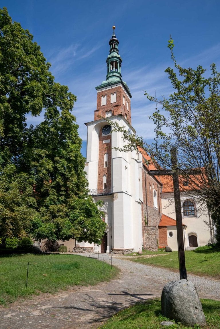 Zdjęcie przedstawia wieżę kościoła rzymskokatolickiegoi pw. św. Leonarda w Lubiniu z białymi elementami architektonicznymi i kamiennym krzyżem na pierwszym planie. W połowie wieży można zauważyć zegar. Kościół otoczony jest zielenią drzew.
