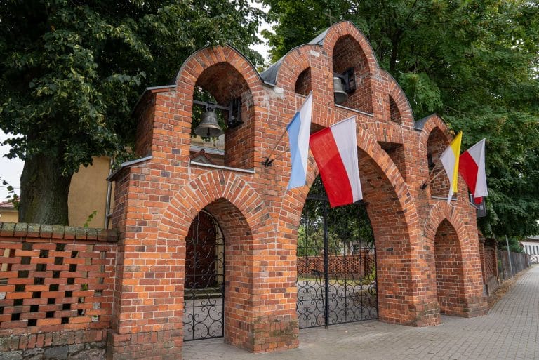 Zdjęcie przedstawia murowaną z czerwonej cegły bramę kościoła rzymskokatolickiego pw. św. Piotra i Pawła w Łuszkowie, składającą się z trzech arkad, w których zawieszone są dzwony. Na bramie powiewają flag. Widoczne elementy muru i drzewa w tle.