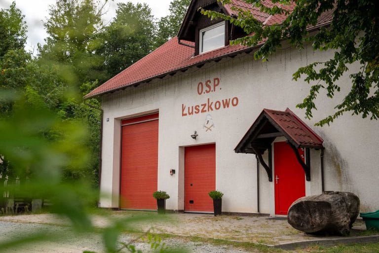 Zdjęcie przedstawia budynek remizy Ochotniczej Straży Pożarnej w Łuszkowie. Biały budynek z dwuspadowym dachem pokrytym czerwoną dachówką. Widoczne dwie czerwone bramy garażowe oraz czerwone drzwi wejściowe z małym zadaszeniem. Za budynkiem widać gęstą zieleń.