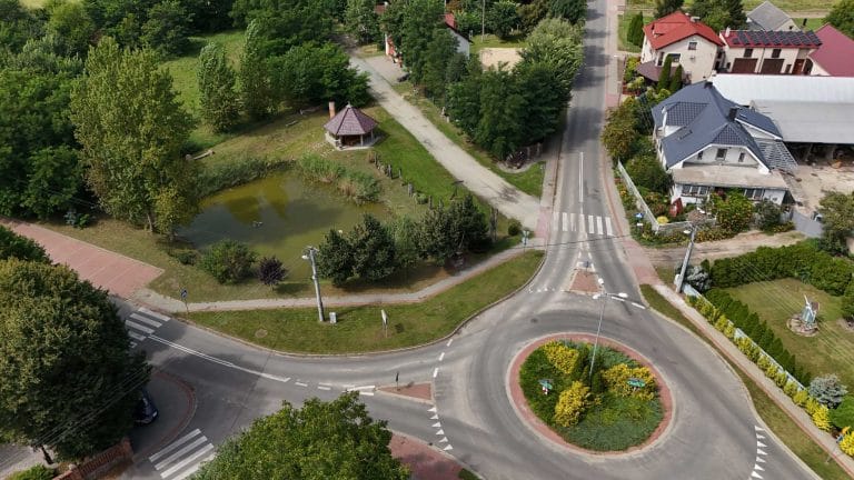 Fotografia lotnicza wsi Łuszkowo przedstawiająca rondo pokryte zielenią. Obok ronda widoczny jest mały staw z atlaną przy brzegu. Wokół widoczny kawałek zabudowy wiejskiej.